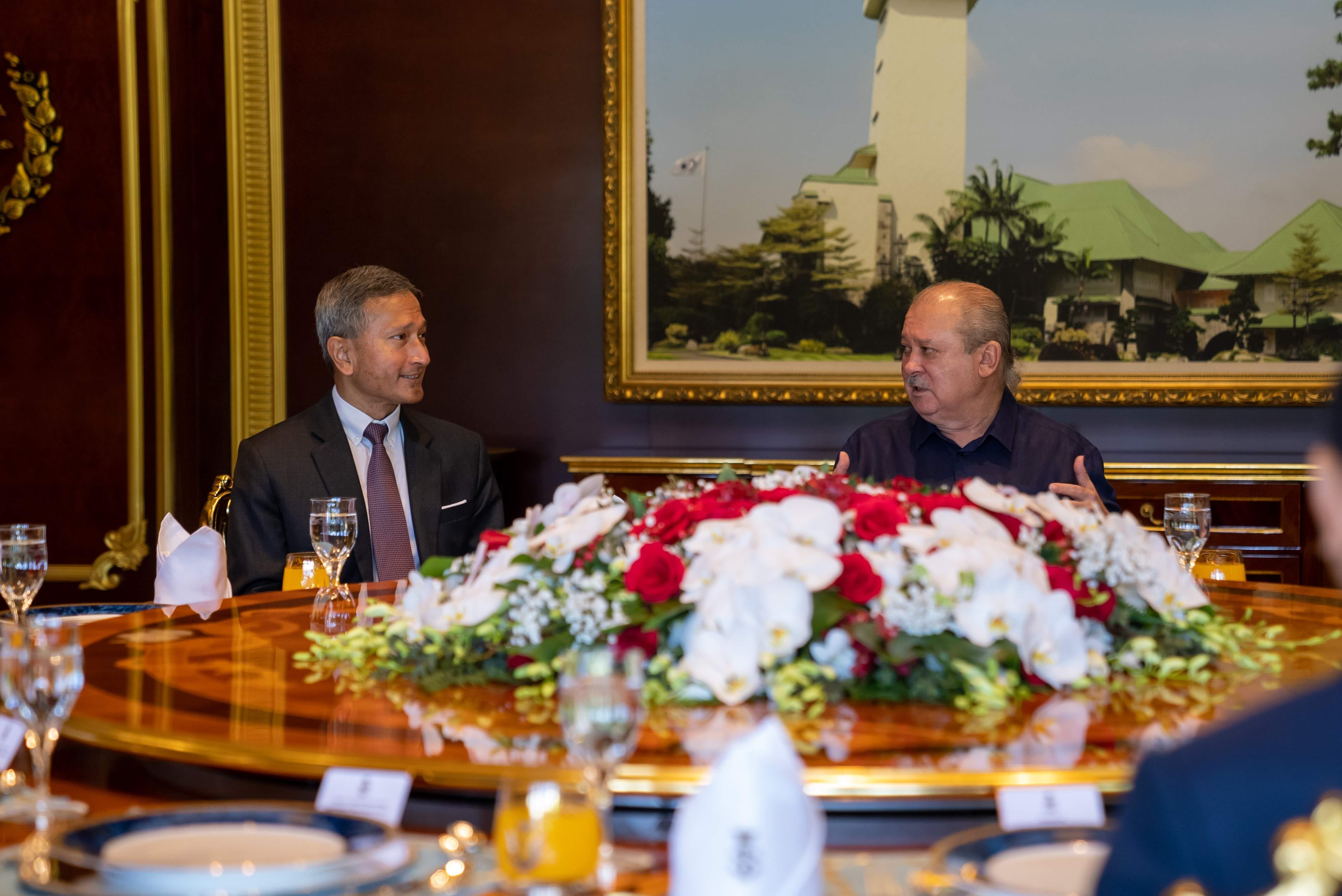 Two men at a table with flowers; a building painting hangs behind them.